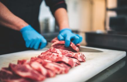closeup view of a young caucasian man cutting raw pork neck with sharp knife. He makes steaks for grill in restaurant kitchen