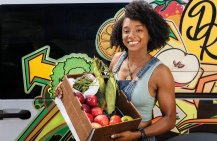 Dazmonique Carr holding a box of produce standing in front of her Deeply Rooted Produce van used for mobile deliveries